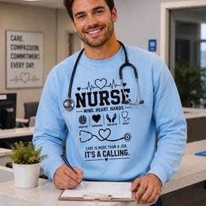 Puede incluir: Una sudadera azul claro de cuello redondo con texto gráfico en negro que dice NURSE, MIND. HEART. HANDS. y CARE IS MORE THAN A JOB, IT'S A CALLING. El diseño incluye iconos de un cerebro, un corazón, unas manos y un estetoscopio con forma de corazón.