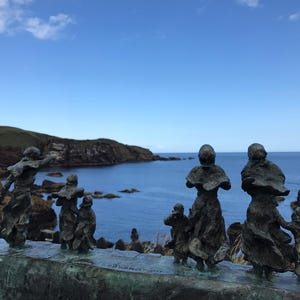 May include: A group of bronze sculptures of people standing on a rock ledge overlooking a body of water. The sculptures are facing the water and appear to be looking out at the horizon. The sky is blue and there are clouds in the distance.