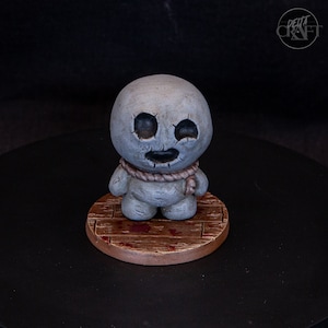 Peut inclure: Figurine grise représentant un personnage ressemblant à une poupée, avec des yeux et une bouche cousus, portant une corde autour du cou. Il se tient sur une plateforme en bois avec des accents rouges. La figurine mesure environ 7.6 cm de haut.