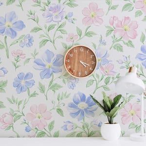 Puede incluir: Papel pintado con un patrón repetitivo de flores azul claro y rosa suave con hojas verdes sobre fondo blanco. Se observa un reloj de pared redondo de madera con números blancos, una lámpara de escritorio blanca y una planta verde en maceta.