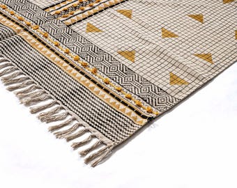 May include: A corner of a beige and black patterned rug with a geometric design. The rug features a fringe on one side and a repeating pattern of black and white stripes, diamonds, and triangles. Yellow accents are incorporated throughout the design.