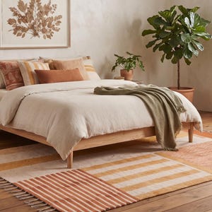 Puede incluir: Una estructura de cama de madera clara con un edredón beige, una manta verde y varios cojines decorativos en tonos naranja y crema. Una alfombra rectangular con rayas naranjas y crema se encuentra en el suelo bajo la estructura de la cama.