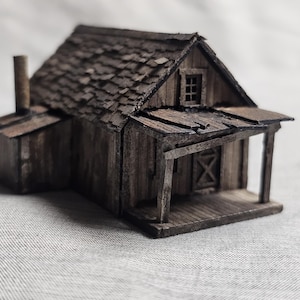 May include: A small wooden model of a rustic cabin with a shingled roof, a porch, and a single window. The model is weathered and has a distressed look.