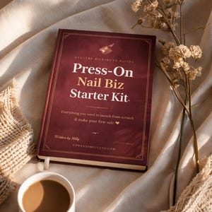 Op de afbeelding: Een bordeauxrood boek met de titel "Press-On Nail Biz Starter Kit" met gouden letters en een kleine illustratie. Het boek ligt open op een beige oppervlak, naast een witte koffiemok en droogbloemen. De tekst op het boek luidt "Everything you need to launch from scratch & make your first sale."