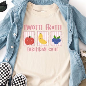 May include: A cream-colored t-shirt with the text "TWOTTI FRUTTI BIRTHDAY CUTIE" and illustrations of an apple, banana, and blueberry. The shirt is paired with a light blue denim shirt, khaki shorts, and black and white checkered slip-on shoes. Black sunglasses are also included.
