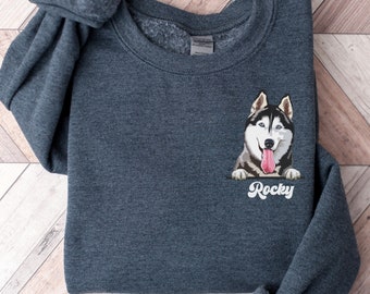 Op de afbeelding: Een donkergrijze sweatshirt met een geborduurd beeld van een husky-hond met blauwe ogen en zijn tong uit. De naam van de hond, "Rocky", is in witte letters geschreven. De sweatshirt ligt op een licht houten oppervlak.