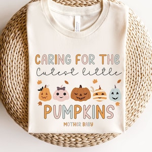 May include: A cream-coloured t-shirt featuring the text "CARING FOR THE Cutest little PUMPKINS" in orange, pink, and blue. The design includes illustrations of pumpkins. The words "MOTHER BABY" are at the bottom of the shirt.