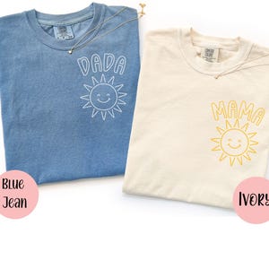 Op de afbeelding: Twee T-shirts, één blauwe jeans en één ivoorkleurig, met de woorden "DADA" en "MAMA" erop gedrukt, elk vergezeld van een lachende zonnegrafiek. De shirts hebben een ronde hals en korte mouwen. Een gouden ketting met een kleine sterhanger is over elk shirt gedrapeerd.