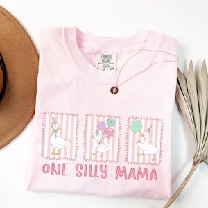 May include: A light pink Comfort Colors t-shirt featuring a graphic of three geese wearing party hats and holding balloons. The text "ONE SILLY MAMA" is printed below the geese. A gold necklace with a pendant is draped on the shirt.