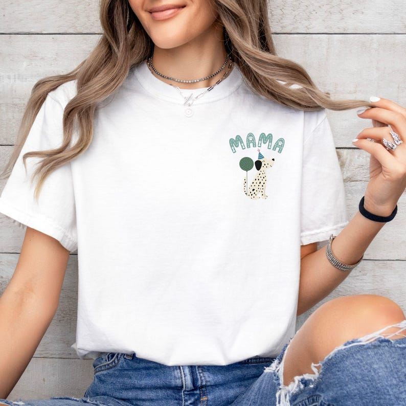 May include: White t-shirt with a graphic of a Dalmatian dog wearing a party hat and holding balloons. The word "MAMA" is above the dog in a light green color. The shirt is made of a soft material.