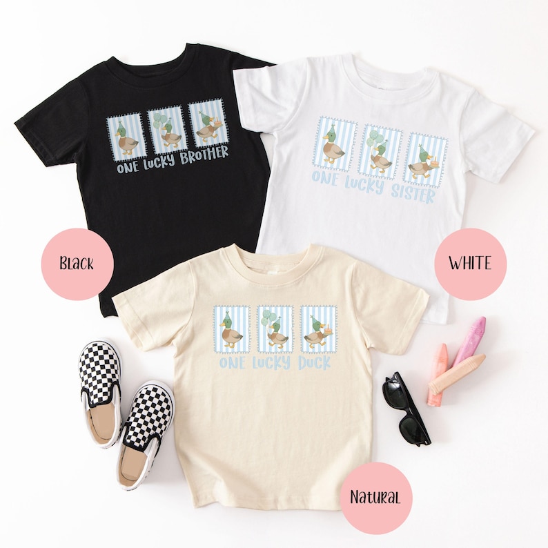 Puede incluir: Tres camisetas de manga corta en colores negro, blanco y natural. Cada camiseta presenta un gr&aacute;fico de pato dentro de un marco a rayas azules y blancas, con el texto "ONE LUCKY BROTHER", "ONE LUCKY SISTER" y "ONE LUCKY DUCK".