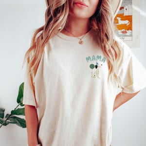 May include: Cream-colored t-shirt featuring the word "MAMA" in light blue bubble letters, with a Dalmatian dog graphic wearing a party hat and holding a balloon. The shirt is a relaxed fit.