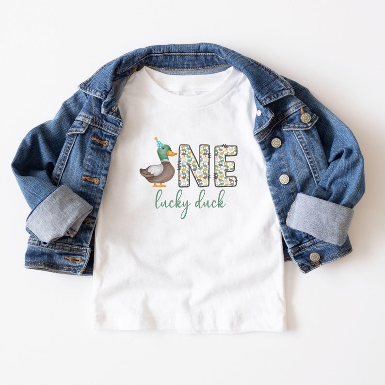 Pode incluir: Camiseta branca com um pato verde e marrom usando um chap&eacute;u de festa azul. O n&uacute;mero "UM" &eacute; escrito em verde e marrom com um padr&atilde;o de pato. O texto "pato da sorte" est&aacute; escrito abaixo do n&uacute;mero.