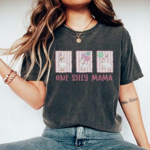 May include: Dark grey t-shirt with a playful design of three geese, each with a party accessory, on pink striped panels. The text "ONE SILLY MAMA" is printed below. The shirt has a vintage, worn appearance, with a relaxed fit.