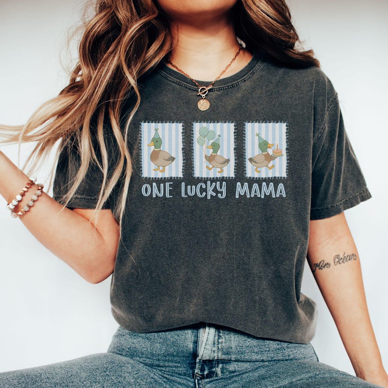 Puede incluir: Camiseta gris oscuro con el texto "ONE LUCKY MAMA". La camiseta presenta tres ilustraciones de patos con sombreros de fiesta y globos. Los patos est&aacute;n sobre un fondo a rayas azules y blancas.