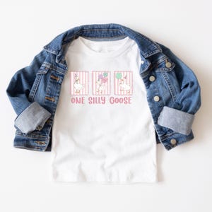 May include: White t-shirt featuring a graphic of three geese with balloons and party hats, with the text "ONE SILLY GOOSE" below. A blue denim jacket is draped over the shirt.