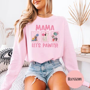 May include: A light pink sweatshirt with the text "MAMA LET'S PAWTY!" and three dog illustrations. The illustrations are in rectangular frames with vertical stripes. The sweatshirt is a long-sleeved crew neck.