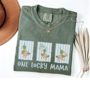 Puede incluir: Una camiseta verde oliva con el texto "ONE LUCKY MAMA" y tres ilustraciones de patos. Cada pato lleva un gorro de fiesta y est&aacute; sobre un fondo a rayas azules y blancas. Tambi&eacute;n se ven un collar dorado y un sombrero marr&oacute;n.