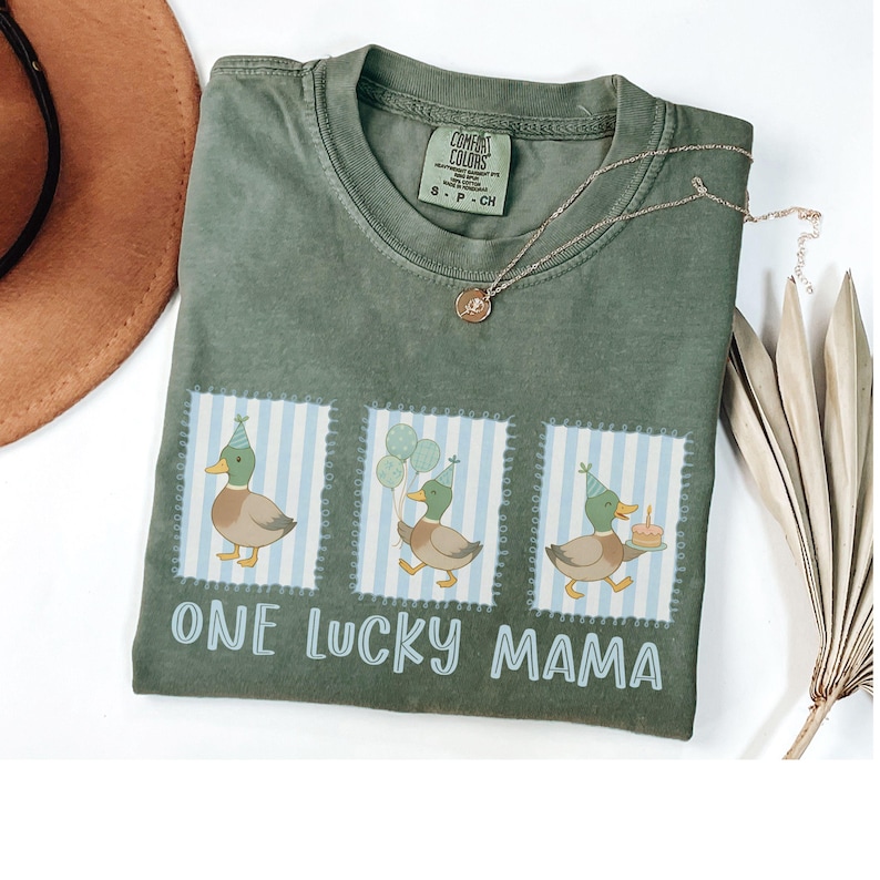Puede incluir: Una camiseta verde oliva con el texto "ONE LUCKY MAMA" y tres ilustraciones de patos. Cada pato lleva un gorro de fiesta y est&aacute; sobre un fondo a rayas azules y blancas. Tambi&eacute;n se ven un collar dorado y un sombrero marr&oacute;n.
