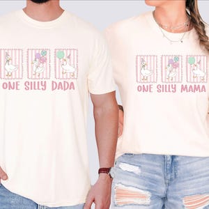 May include: Cream-coloured t-shirts with a whimsical design featuring a goose wearing a party hat and holding balloons. The text "ONE SILLY DADA" and "ONE SILLY MAMA" are printed in pink. The design is framed with a pink border.