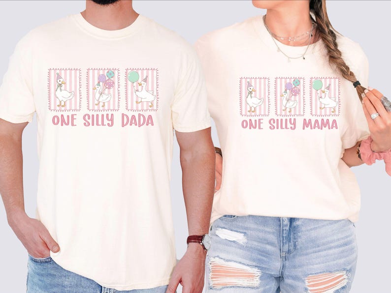May include: Cream-coloured t-shirts with a whimsical design featuring a goose wearing a party hat and holding balloons. The text "ONE SILLY DADA" and "ONE SILLY MAMA" are printed in pink. The design is framed with a pink border.