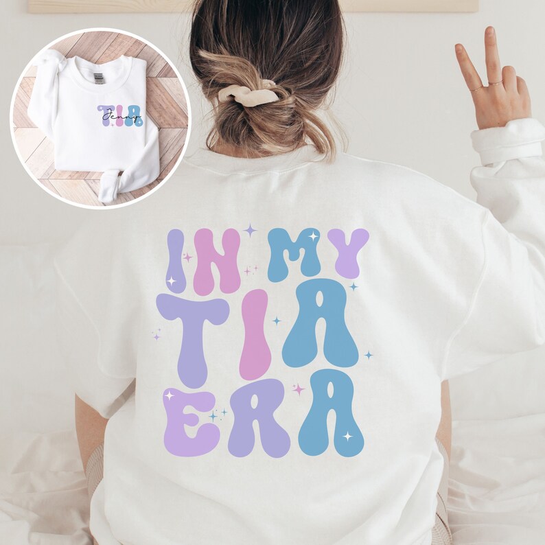 May include: White sweatshirt with the phrase "IN MY TIA ERA" in large, colorful, retro-style lettering on the back. The front of the sweatshirt has the word "TIA" in a smaller, matching font. The design includes pastel blue, purple, and pink colors.
