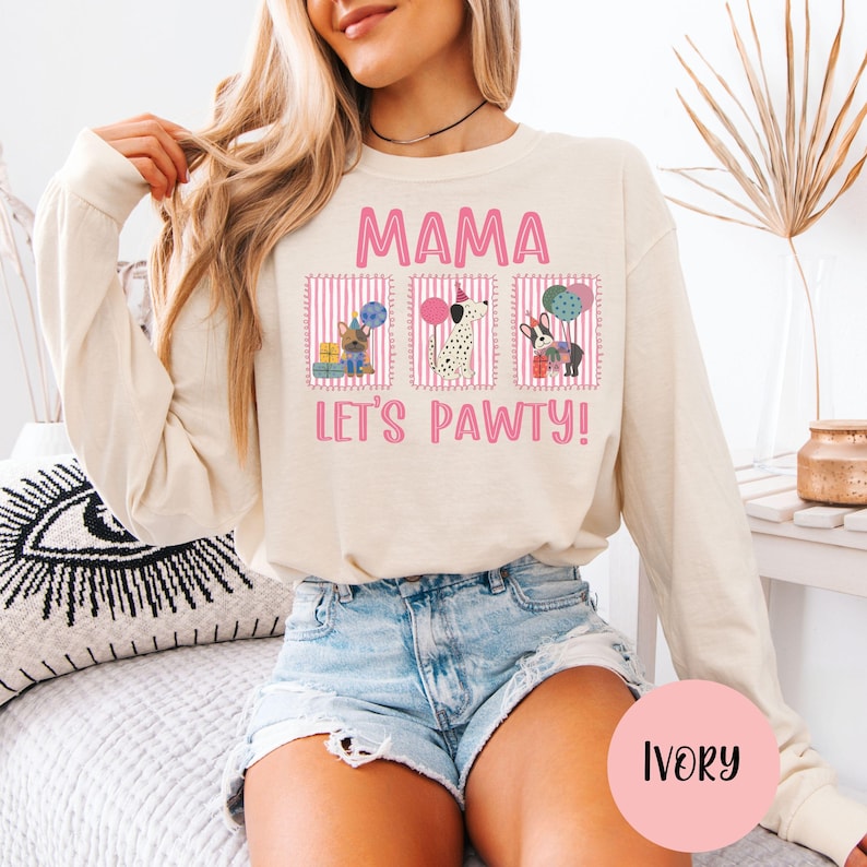 May include: Ivory long-sleeve sweatshirt with the text "MAMA LET'S PAWTY!" in pink. The graphic features three dog illustrations with party hats and balloons. The sweatshirt is a neutral color and has a relaxed fit.