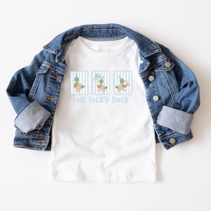 Puede incluir: Camiseta blanca con una chaqueta vaquera azul. La camiseta presenta tres ilustraciones de patos dentro de marcos a rayas azules, cada uno con un gorro de fiesta o globos. El texto "ONE LUCKY DUCK" est&aacute; impreso debajo.