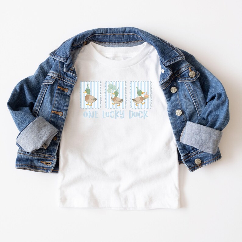 Puede incluir: Camiseta blanca con una chaqueta vaquera azul. La camiseta presenta tres ilustraciones de patos dentro de marcos a rayas azules, cada uno con un gorro de fiesta o globos. El texto "ONE LUCKY DUCK" est&aacute; impreso debajo.