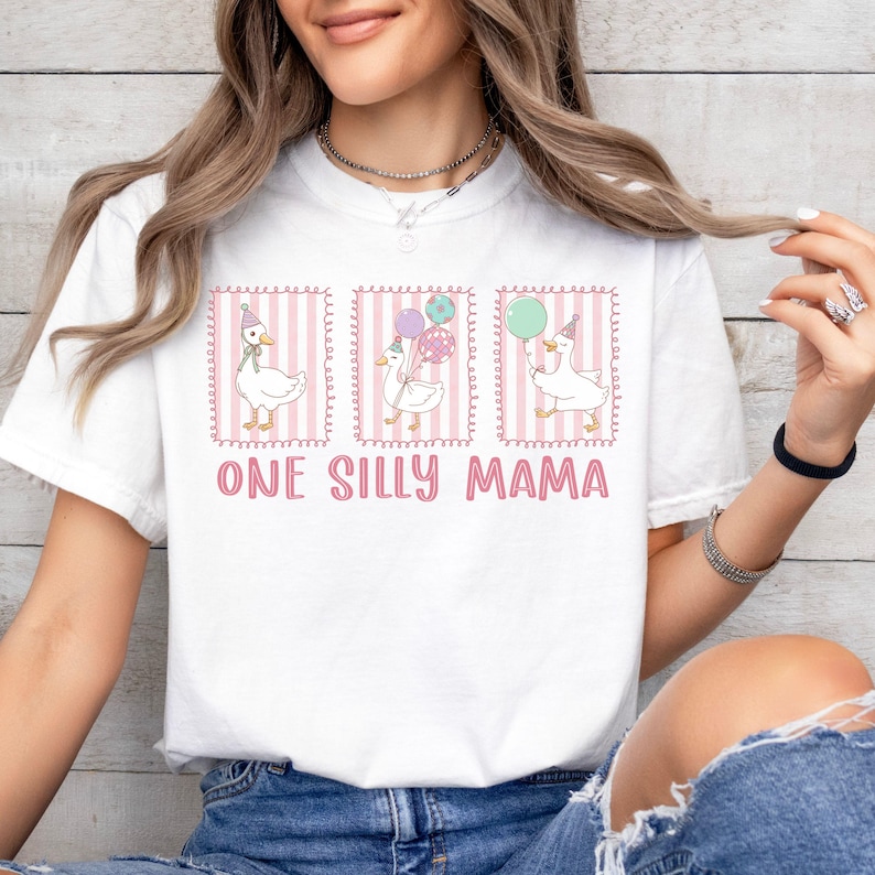 May include: White t-shirt with three framed illustrations of cartoon geese wearing party hats and holding balloons. The text "ONE SILLY MAMA" is printed in pink below the illustrations. The shirt is worn by a person.