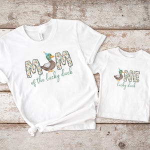 Pode incluir: Duas camisetas brancas com um desenho de pato e texto. A camiseta maior diz "MOM of the lucky duck" e a menor diz "ONE lucky duck".