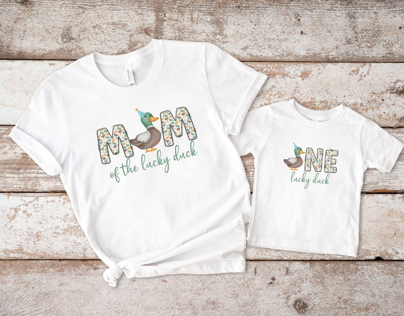Pode incluir: Duas camisetas brancas com um desenho de pato e texto. A camiseta maior diz "MOM of the lucky duck" e a menor diz "ONE lucky duck".