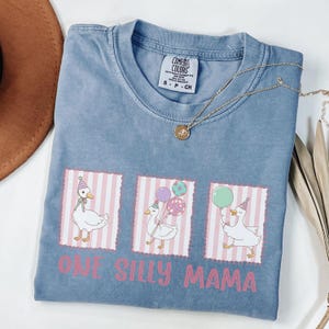 May include: A blue Comfort Colors t-shirt with three cartoon geese wearing party hats and holding balloons. The text "ONE SILLY MAMA" is printed in pink. A gold necklace is draped across the shirt. The shirt is size S-CH.