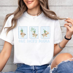 Puede incluir: Camiseta blanca con un gr&aacute;fico de tres patos con sombreros de fiesta, cada uno en un marco a rayas azules. Los patos sostienen globos y un pastel. El texto "ONE LUCKY MAMA" est&aacute; impreso en azul claro.