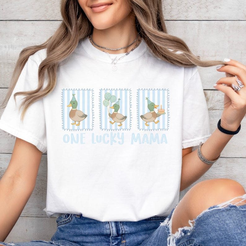 Puede incluir: Camiseta blanca con un gr&aacute;fico de tres patos con sombreros de fiesta, cada uno en un marco a rayas azules. Los patos sostienen globos y un pastel. El texto "ONE LUCKY MAMA" est&aacute; impreso en azul claro.