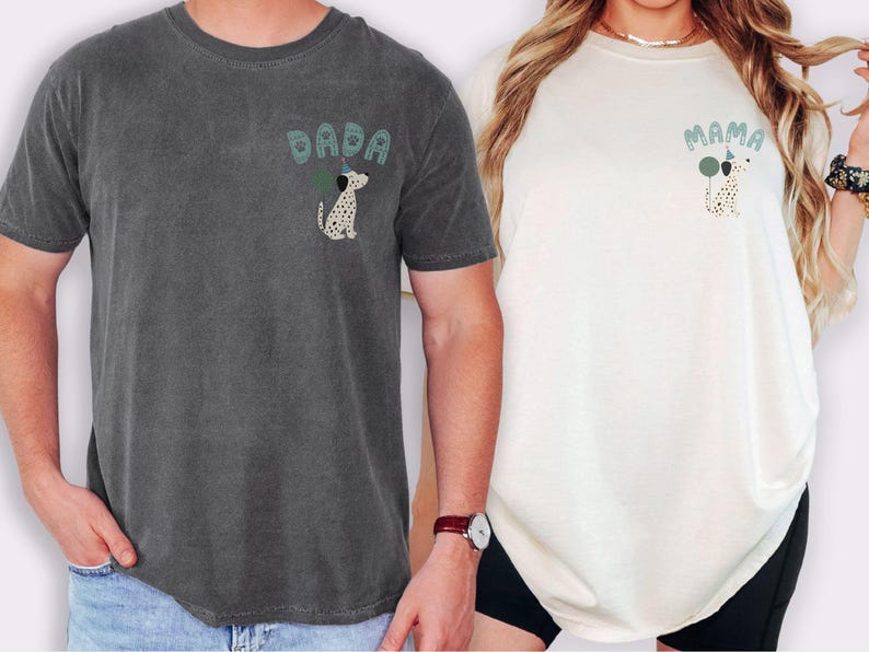 May include: Two t-shirts, one charcoal gray and one off-white, each with a graphic of a Dalmatian dog holding a balloon. The charcoal shirt says "DADA" and the off-white shirt says "MAMA" in green lettering. Both shirts are short-sleeved.