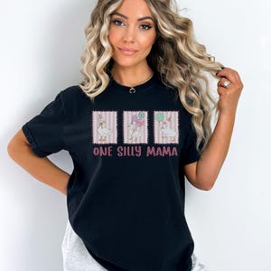 May include: Black t-shirt featuring a graphic of three geese with balloons. The text "ONE SILLY MAMA" is printed in pink below the geese. The shirt is a short-sleeved, casual style.