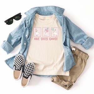May include: A cream-coloured t-shirt with the text "ONE SILLY GOOSE" and three cartoon geese with party hats and balloons. The shirt is paired with a light blue denim jacket, khaki shorts, black and white checkered slip-on shoes, and black sunglasses.