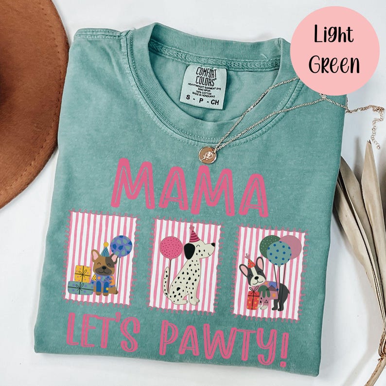 May include: Light green t-shirt with the words "MAMA" and "LET'S PAWTY!" in pink. The shirt features three dog illustrations with party hats and balloons. A silver necklace is draped across the shirt.