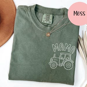 Peut inclure: Un t-shirt vert mousse avec un dessin blanc d'un tracteur et le mot "MAMA". Le t-shirt a un col rond et des manches courtes. Un collier doré est drapé sur le t-shirt. Le mot "MOSS" est dans un cercle rose.