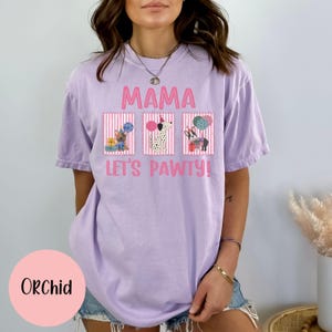 May include: Orchid-colored t-shirt with the text "MAMA LET'S PAWTY!" and three dog illustrations. The illustrations include a French Bulldog, a Dalmatian, and a dog with a gift. The shirt is a relaxed fit.
