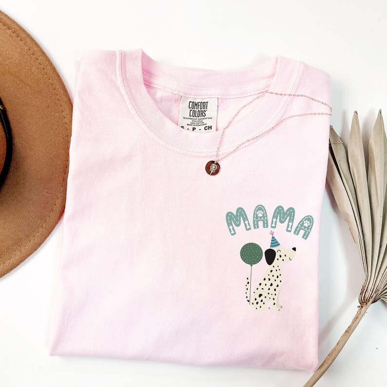 May include: A light pink Comfort Colors t-shirt with the word "MAMA" in teal letters and a Dalmatian dog graphic holding a green balloon. A rose gold necklace is draped across the shirt.