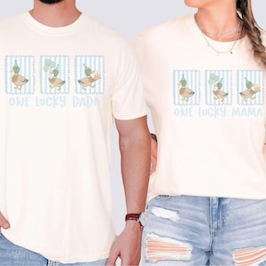 Puede incluir: Camisetas color crema con un gr&aacute;fico de pato y el texto "ONE LUCKY DADA" y "ONE LUCKY MAMA". El gr&aacute;fico del pato est&aacute; en un marco a rayas azules y blancas, con los patos usando sombreros de fiesta y sosteniendo globos y un pastel.