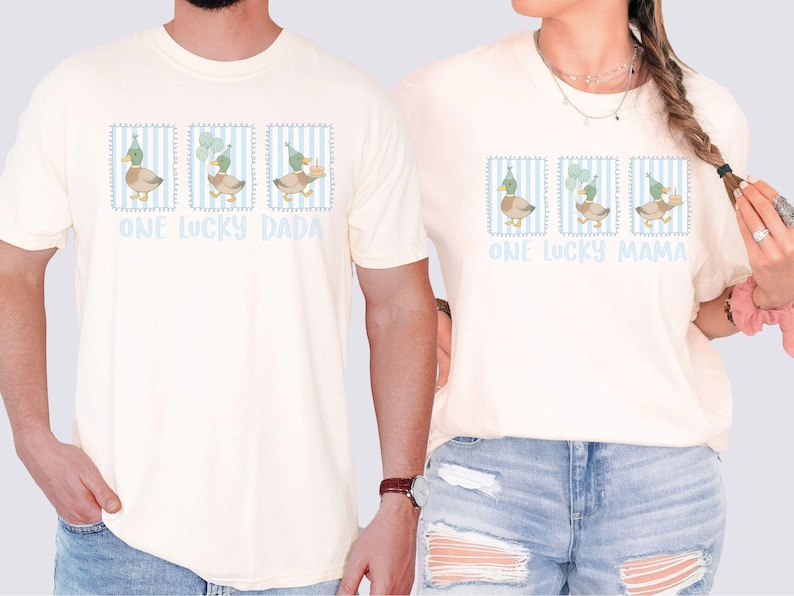 Puede incluir: Camisetas color crema con un gr&aacute;fico de pato y el texto "ONE LUCKY DADA" y "ONE LUCKY MAMA". El gr&aacute;fico del pato est&aacute; en un marco a rayas azules y blancas, con los patos usando sombreros de fiesta y sosteniendo globos y un pastel.