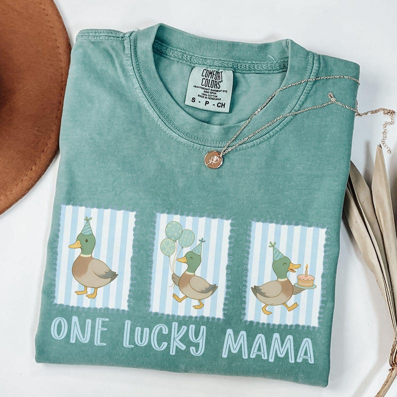 Puede incluir: Una camiseta Comfort Colors color verde azulado con un gr&aacute;fico de tres patos con sombreros de fiesta, globos y un pastel. El texto "ONE LUCKY MAMA" est&aacute; impreso debajo. Un collar plateado con un colgante est&aacute; sobre la camiseta.
