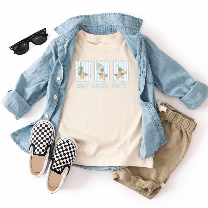Puede incluir: Una camiseta color crema con el texto "ONE LUCKY DUCK" y tres ilustraciones de patos. La camiseta se combina con una camisa vaquera azul claro, pantalones cortos color caqui, zapatos sin cordones a cuadros blancos y negros y gafas de sol negras.