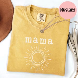 Op de afbeelding: Mosterdkleurig T-shirt met het woord "mama" in witte blokletters boven een wit zonnesterontwerp. Een zilveren ketting met een kleine hanger rust op het shirt. Het shirt is van het merk Comfort Colors.