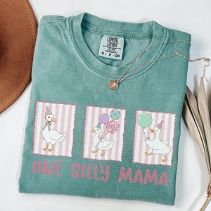 May include: A teal Comfort Colors t-shirt with a graphic of three geese wearing party hats and holding balloons. The text "ONE SILLY MAMA" is printed in pink below the geese. A silver necklace with a pendant is draped across the shirt.