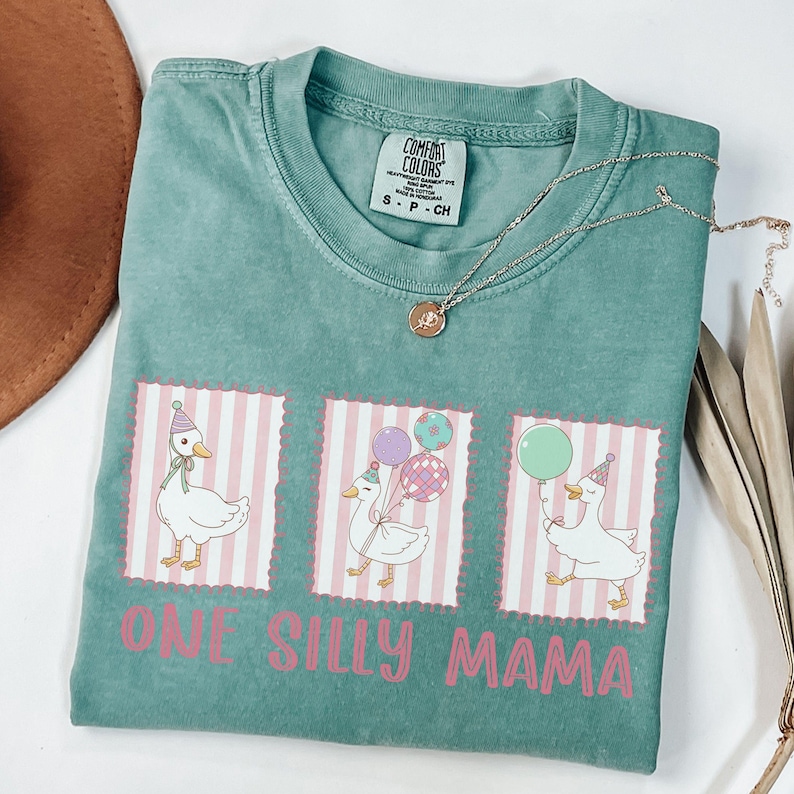 May include: A teal Comfort Colors t-shirt with a graphic of three geese wearing party hats and holding balloons. The text "ONE SILLY MAMA" is printed in pink below the geese. A silver necklace with a pendant is draped across the shirt.