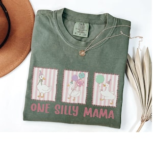 May include: Sage green t-shirt featuring the text "ONE SILLY MAMA" and three cartoon geese wearing party hats and holding balloons. The geese are in pink and white striped frames. A gold necklace is draped across the shirt.